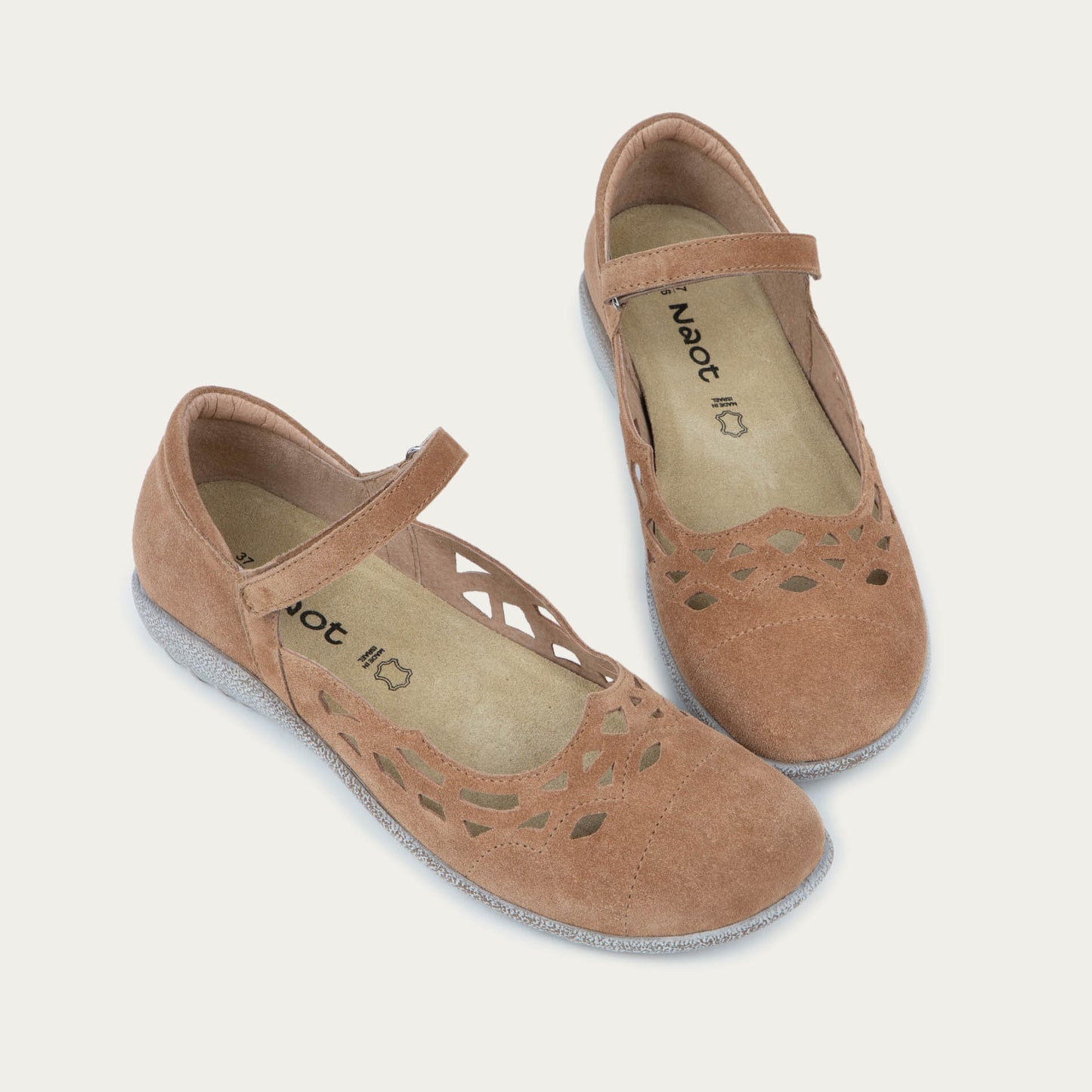 Pair of Tan suede Mary Jane shoes with cut-out patterns, Velcro straps, and textured gray soles, set against a plain white background. Casual and stylish.