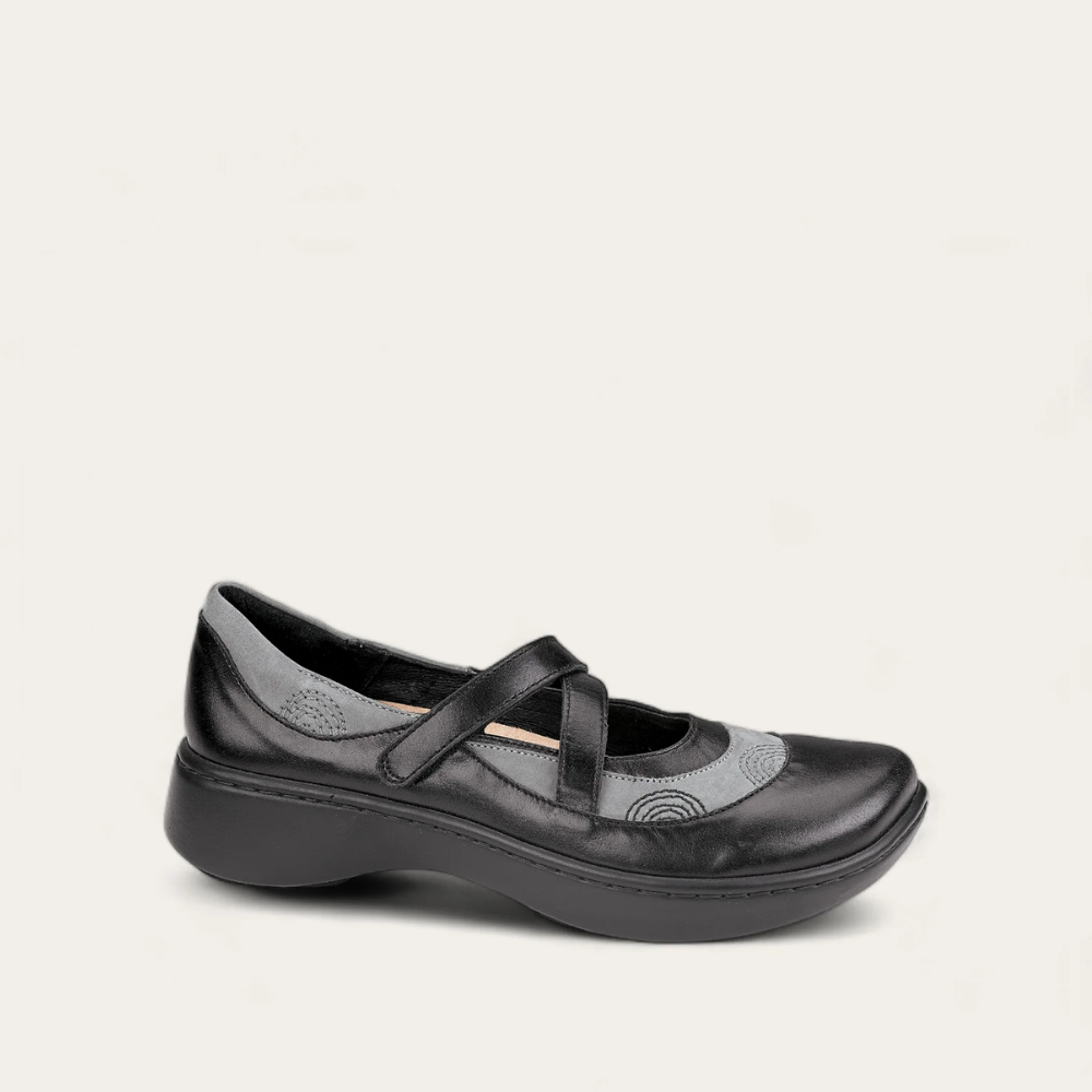A black Mary Jane flat shoe with a hook and loop closure and contrasting leathers. It features gores at the instep for adjustability and stretch.