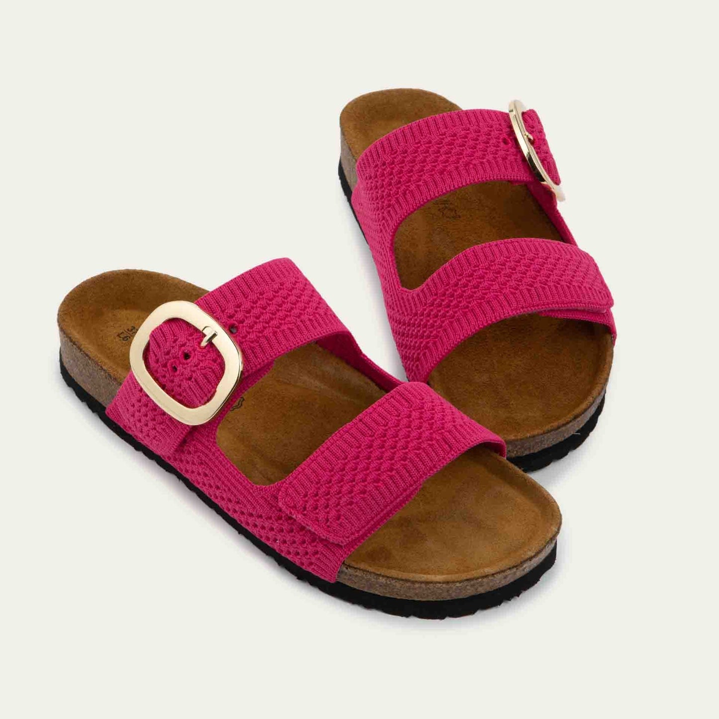 Pair of pink slip-on sandals with two adjustable knit straps and a large gold buckle. It has a cork footbed and a black sole.