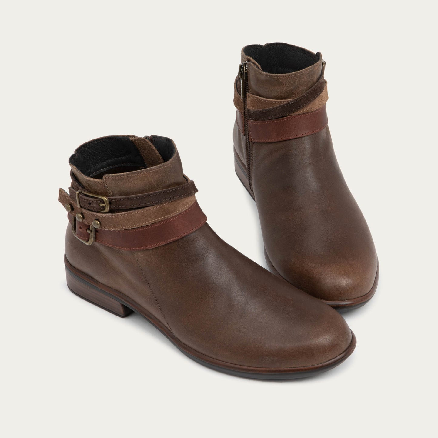 A pair of brown leather ankle boots with two decorative straps and brass buckles. These boots has a low heel and a smooth, polished finish, conveying a stylish look.