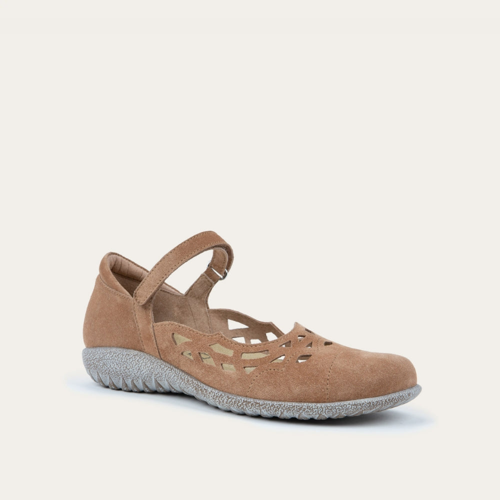 Angled view of a Tan suede Mary Jane shoe with cut-out patterns, a Velcro strap, and a textured gray sole, set against a plain white background. Casual and stylish.