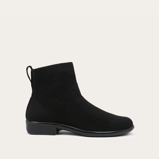 Profile view of a black ankle boot featuring a textured knit upper, a pull tab at the heel, and a low, flat black sole.
