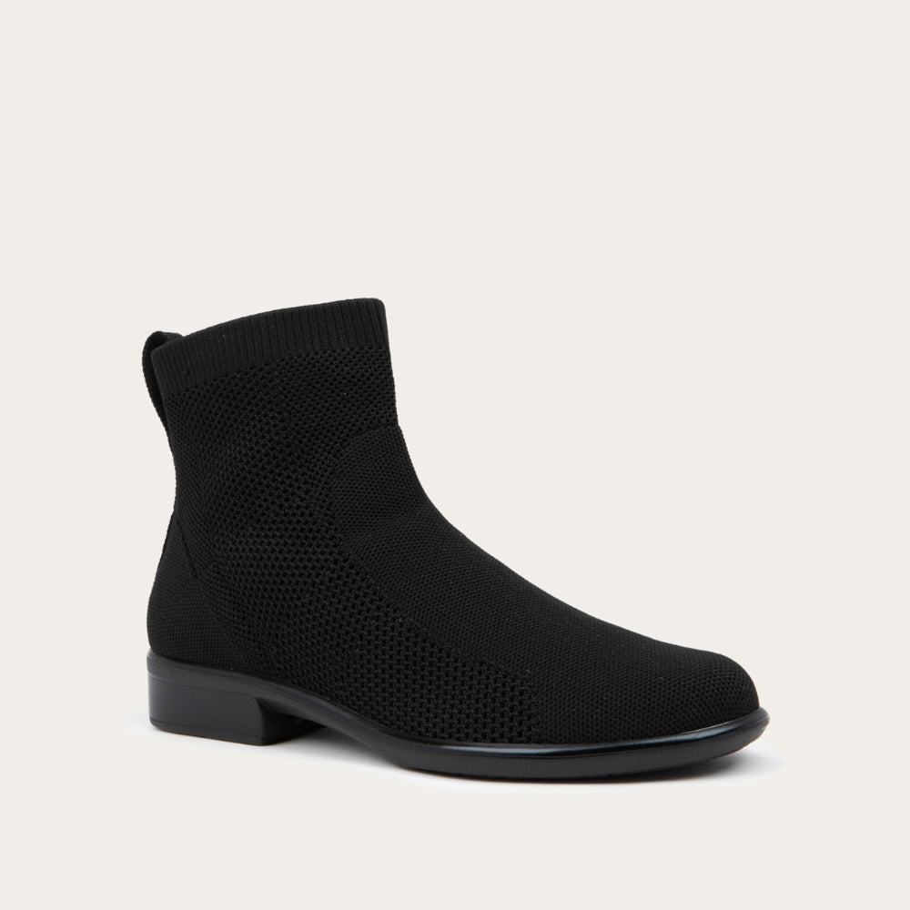 Angled profile view of a black ankle boot featuring a textured knit upper, a pull tab at the heel, and a low, flat black sole.