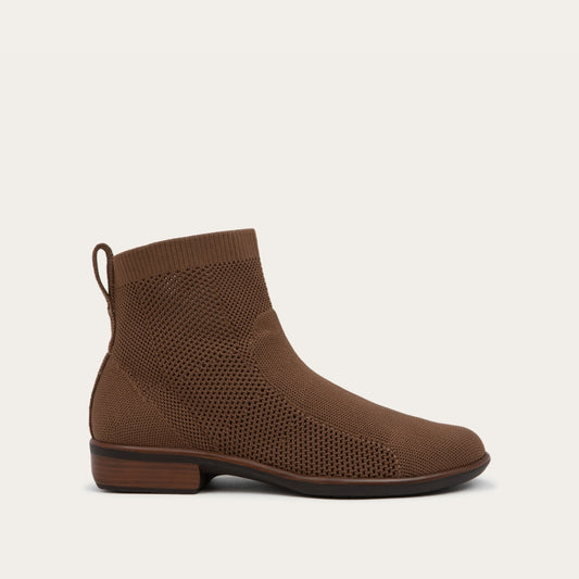 Profile view of a brown ankle boot featuring a textured knit upper, a pull tab at the heel, and a low, flat brown sole.