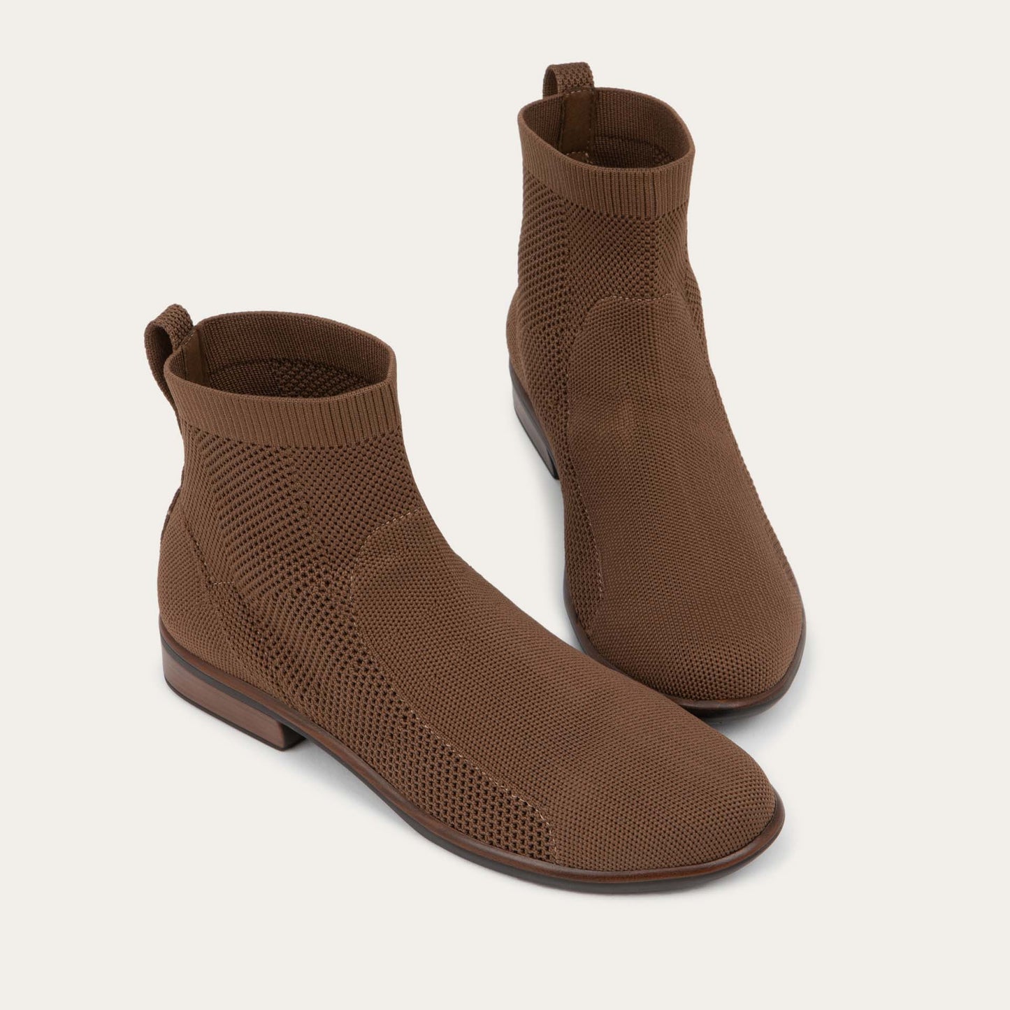 A pair of of brown ankle boots featuring a textured knit uppers, pull tabs at the heels, and low, flat brown soles.