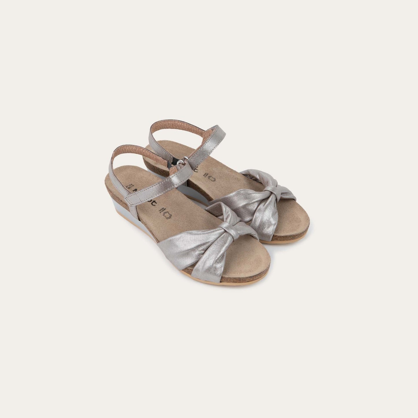 A pair of  silver wedge sandals with an ankle strap and twisted front strap, featuring a cork footbed and wedge heel. 