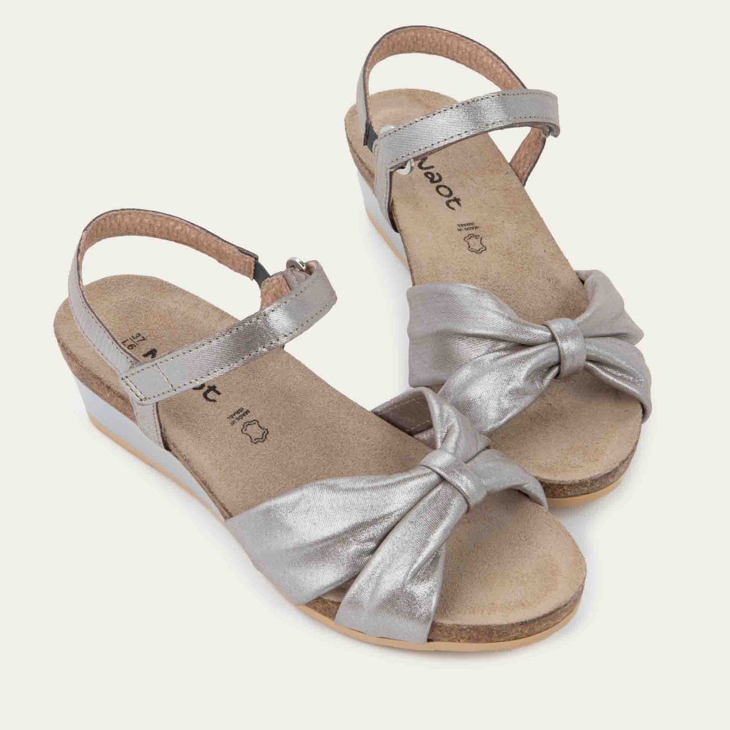 A pair of  silver wedge sandals with an ankle strap and twisted front strap, featuring a cork footbed and wedge heel. 