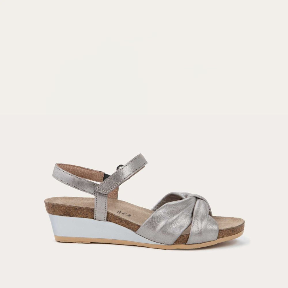 Profile view of a silver wedge sandal with an ankle strap and twisted front strap, featuring a cork footbed and wedge heel. 