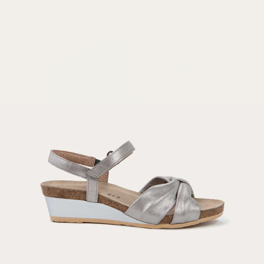 Profile view of a silver wedge sandal with an ankle strap and twisted front strap, featuring a cork footbed and wedge heel. 