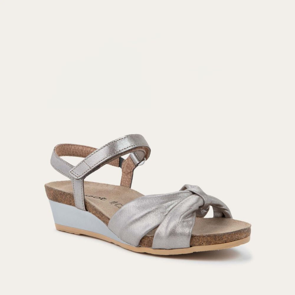 Angled profile view of a silver wedge sandal with an ankle strap and twisted front strap, featuring a cork footbed and wedge heel. 