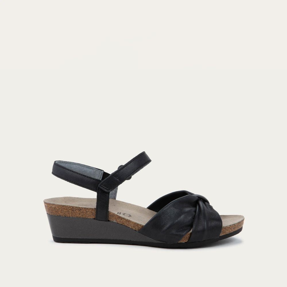 Profile view of a black wedge sandal with an ankle strap and twisted front strap, featuring a cork footbed and wedge heel. 