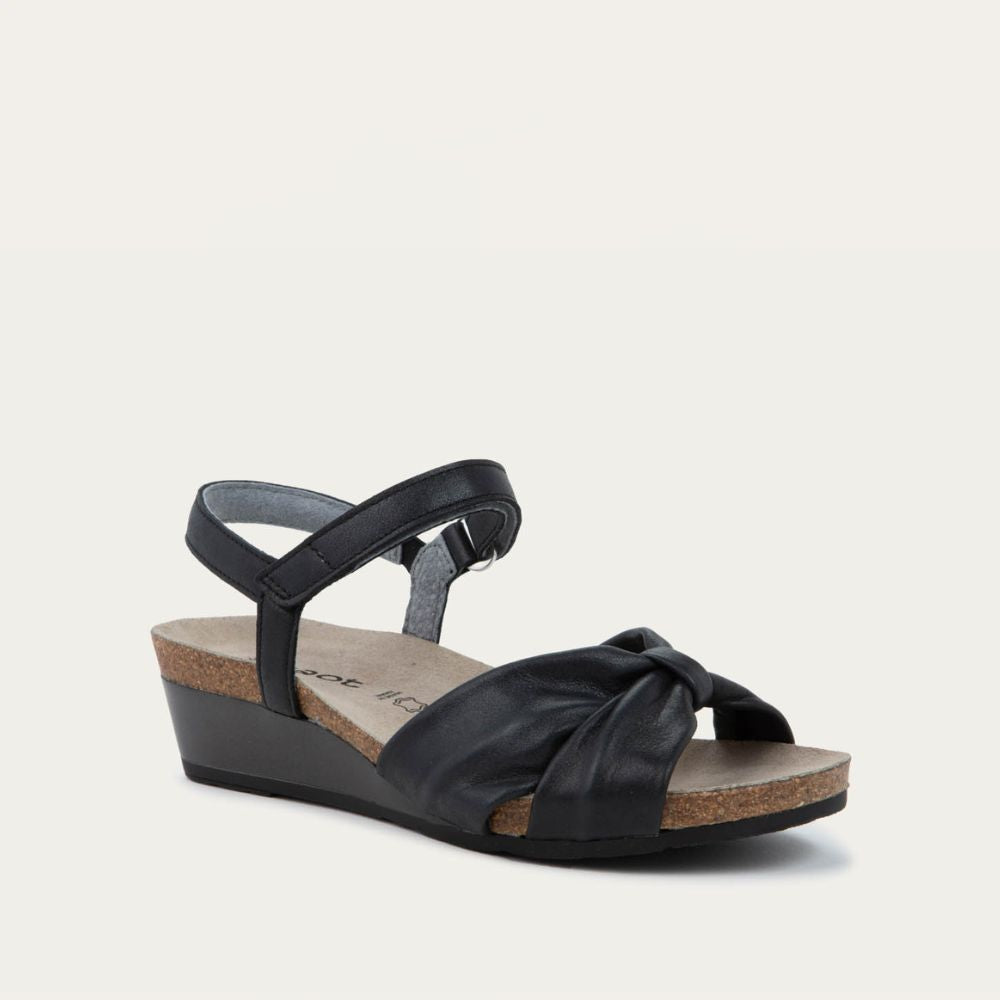 Angled profile view of a black wedge sandal with an ankle strap and twisted front strap, featuring a cork footbed and wedge heel. 