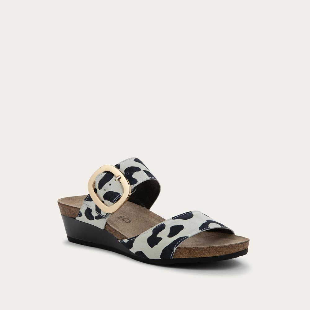 Angled profile view of a  black and white leaopard-print slip on wedge sandal with two straps and a large gold buckle. 