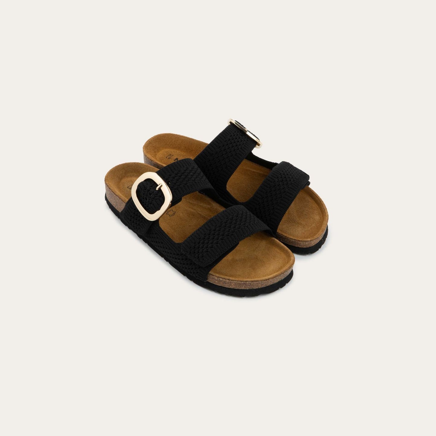 Pair of black slip-on sandals with two adjustable knit straps and a large gold buckle. It has a cork footbed and a black sole.