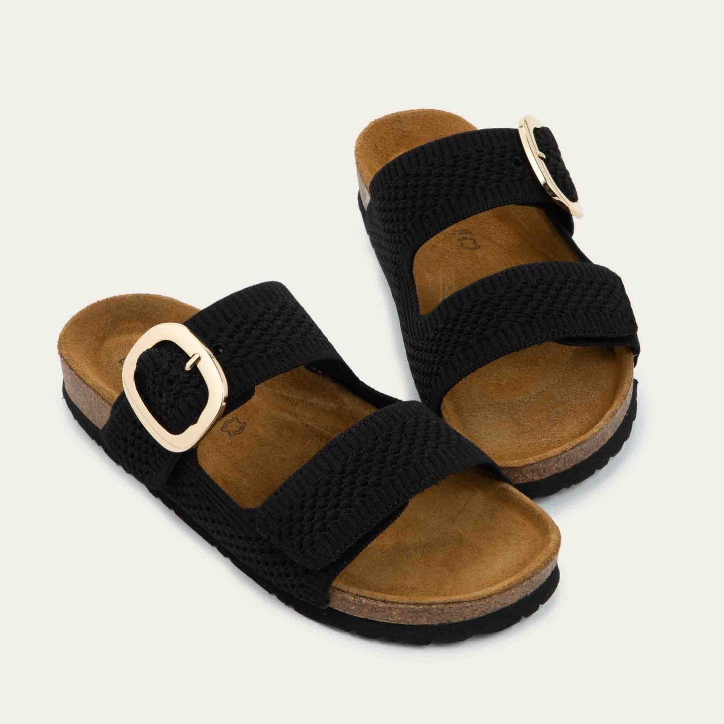 Pair of black slip-on sandals with two adjustable knit straps and a large gold buckle. It has a cork footbed and a black sole.