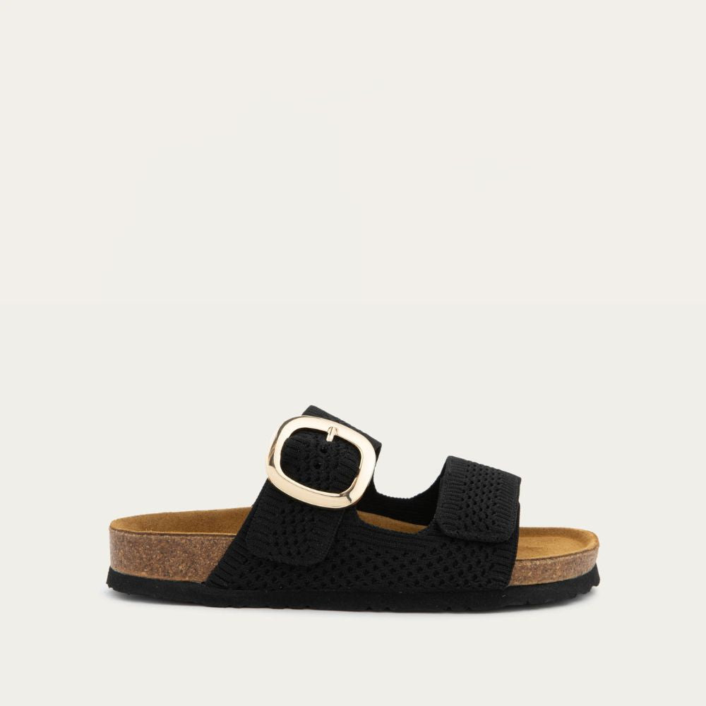 Profile view of a black slip-on sandal with two adjustable knit straps and a large gold buckle. It has a cork footbed and a black sole.