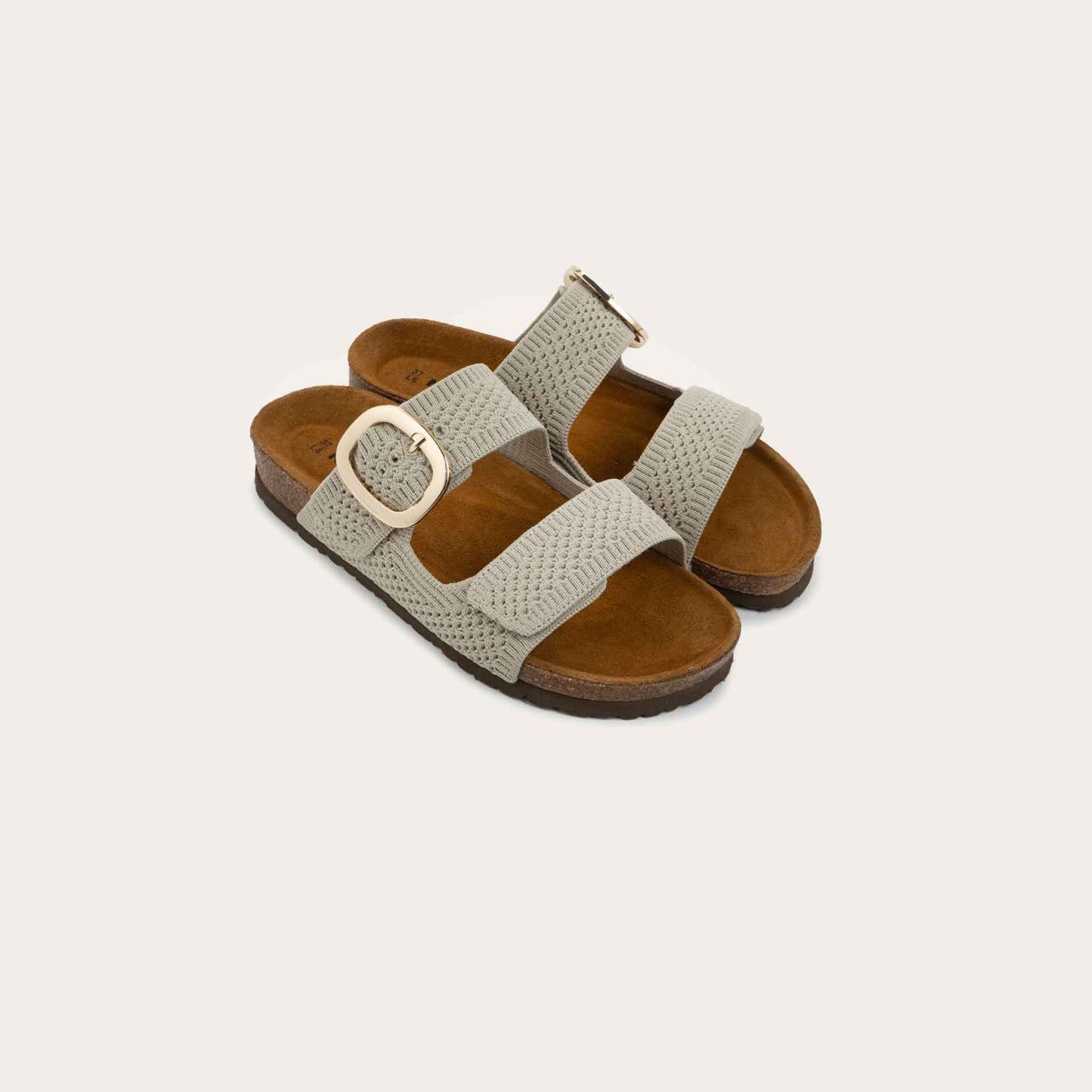 Pair of beige slip-on sandals with two adjustable knit straps and a large gold buckle. It has a cork footbed and a black sole.