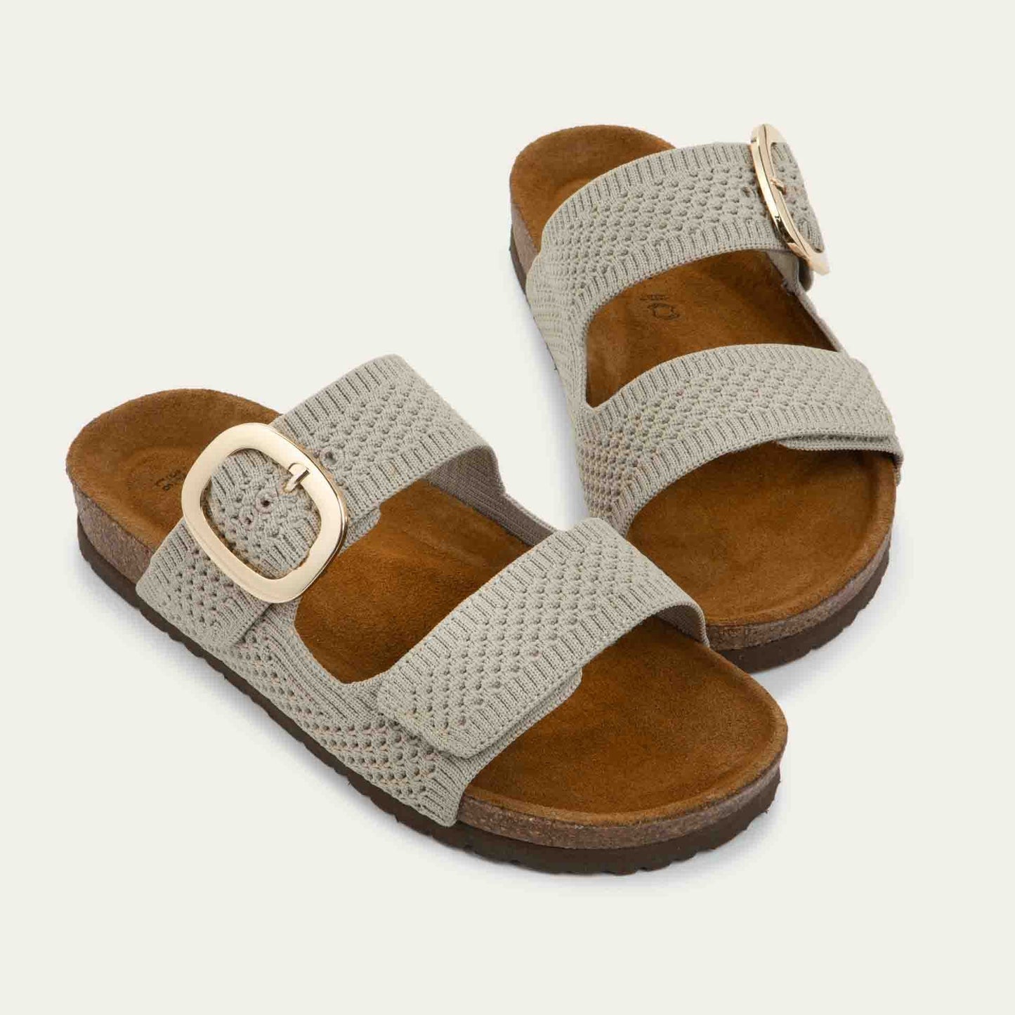 Pair of beige  slip-on sandals with two adjustable knit straps and a large gold buckle. It has a cork footbed and a black sole.