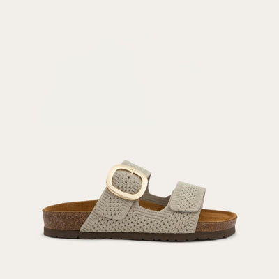 Profile view of a beige slip-on sandal with two adjustable knit straps and a large gold buckle. It has a cork footbed and a black sole.