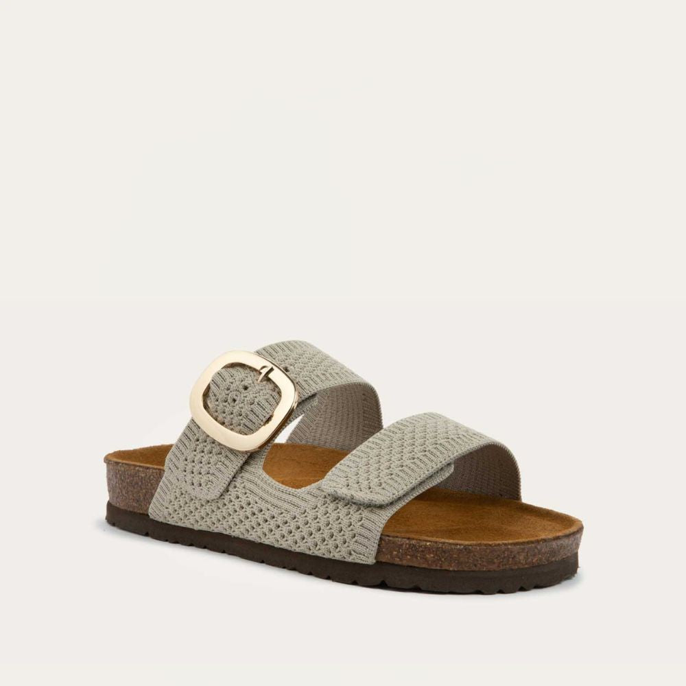Angled profile view of a beige  slip-on sandal with two adjustable knit straps and a large gold buckle. It has a cork footbed and a black sole.