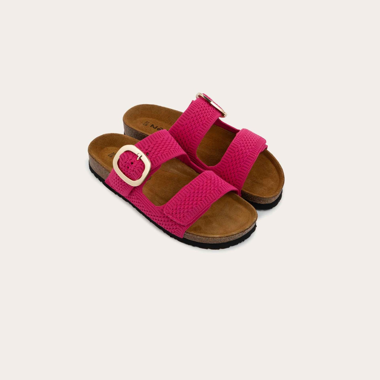 Pair of pink slip-on sandals with two adjustable knit straps and a large gold buckle. It has a cork footbed and a black sole.
