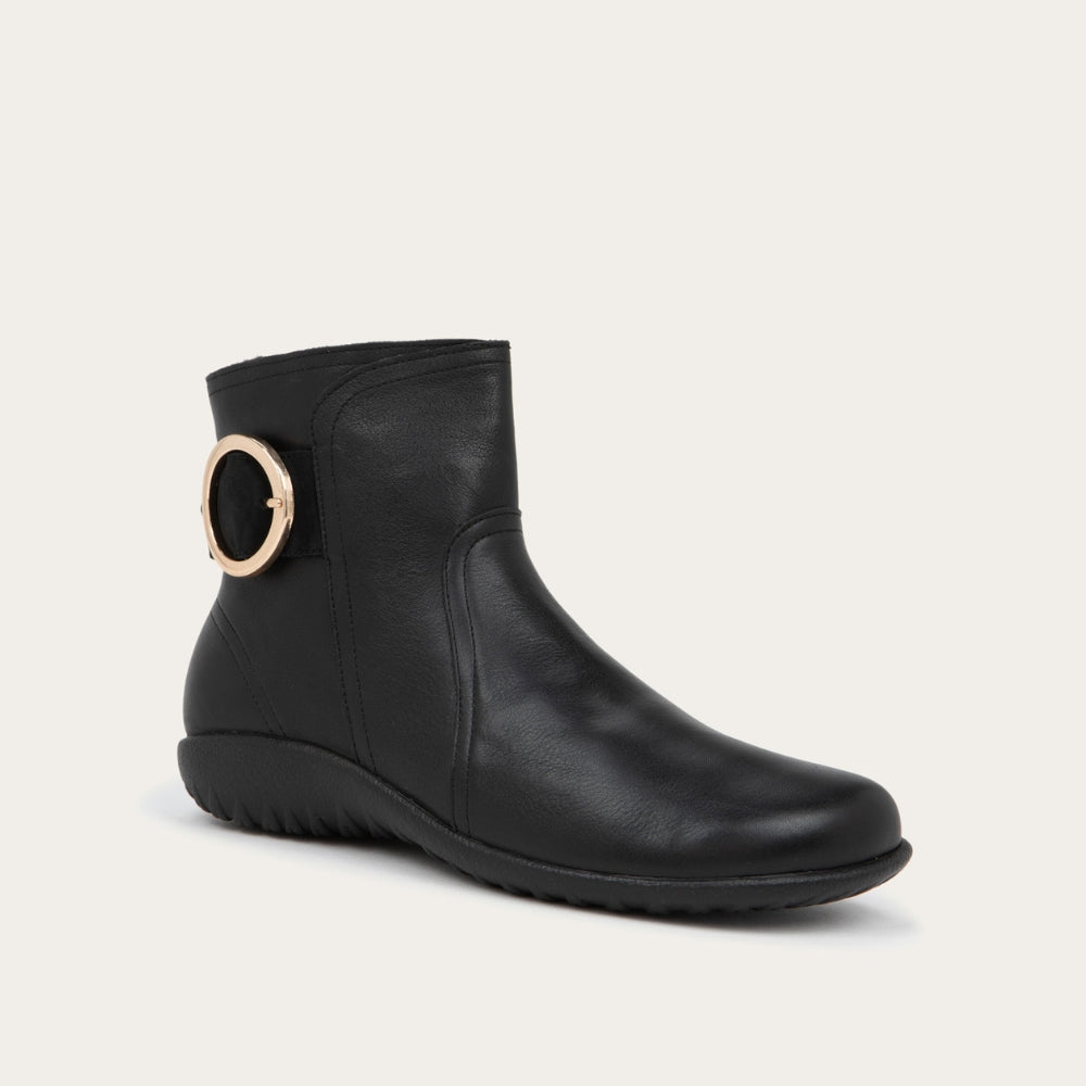 Angled profile view of a black boot featuring an accent strap with round buckle in the back of the shoe on a dark sole.