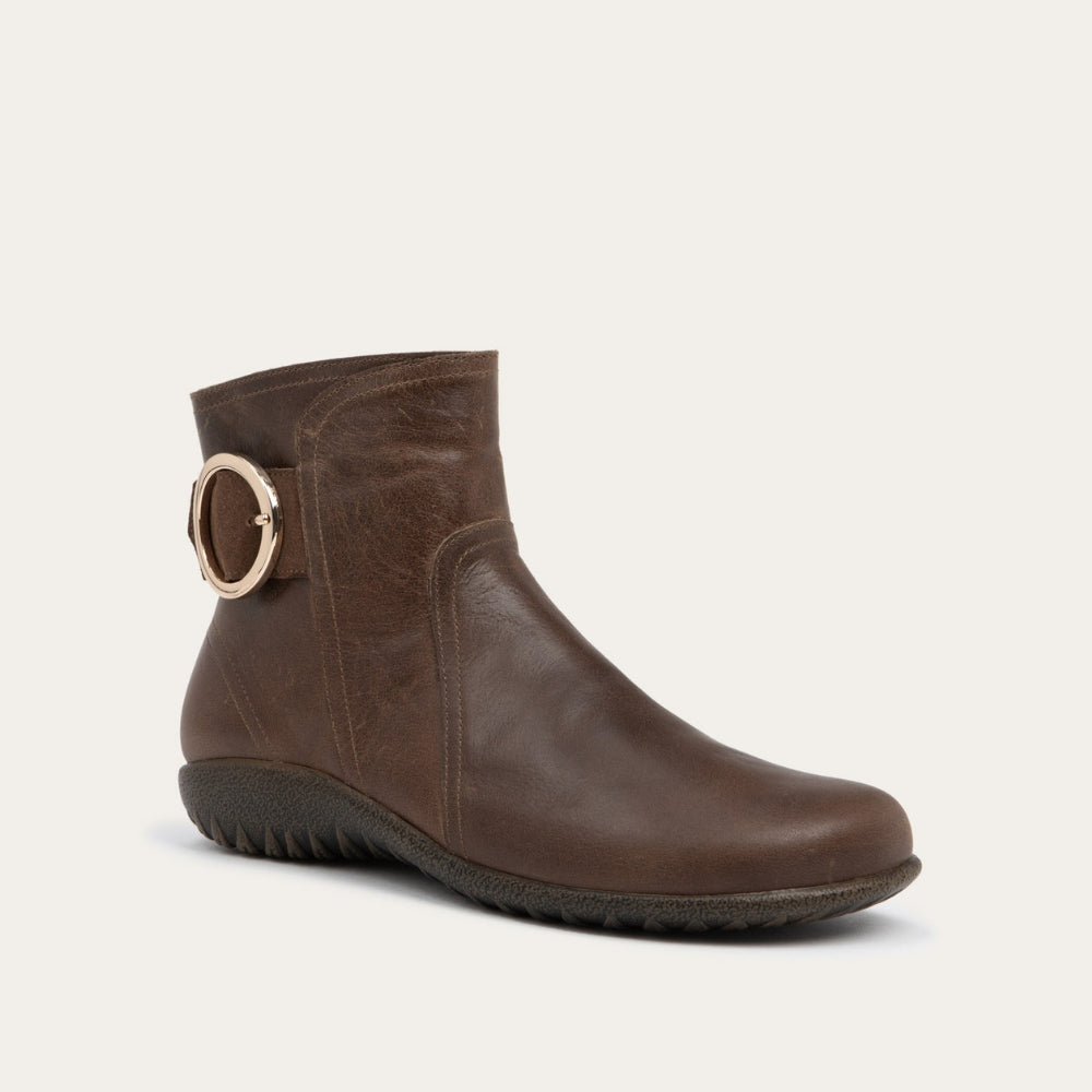 Angled profile view of a brown boot featuring an accent strap with round buckle in the back of the shoe on a dark sole.