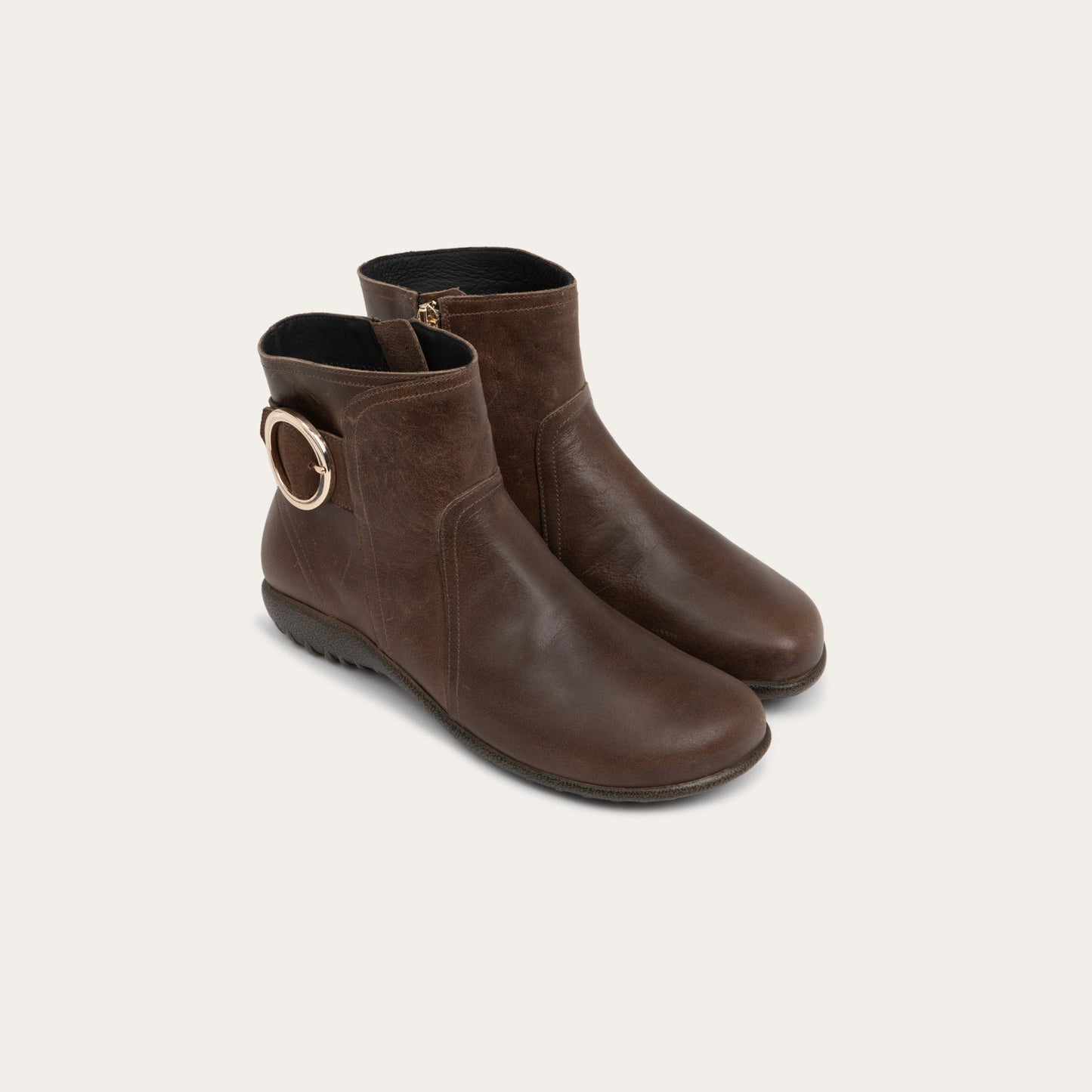 Pair of brown boots featuring an accent strap with round buckle in the back of the shoe on a dark sole.