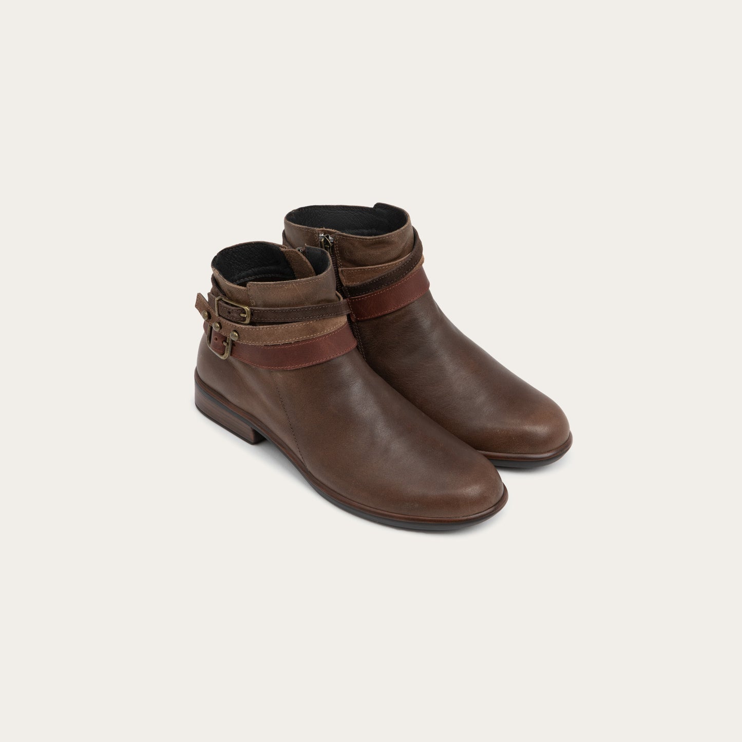 A pair of brown leather ankle boots with two decorative straps and brass buckles. These boots has a low heel and a smooth, polished finish, conveying a stylish look.