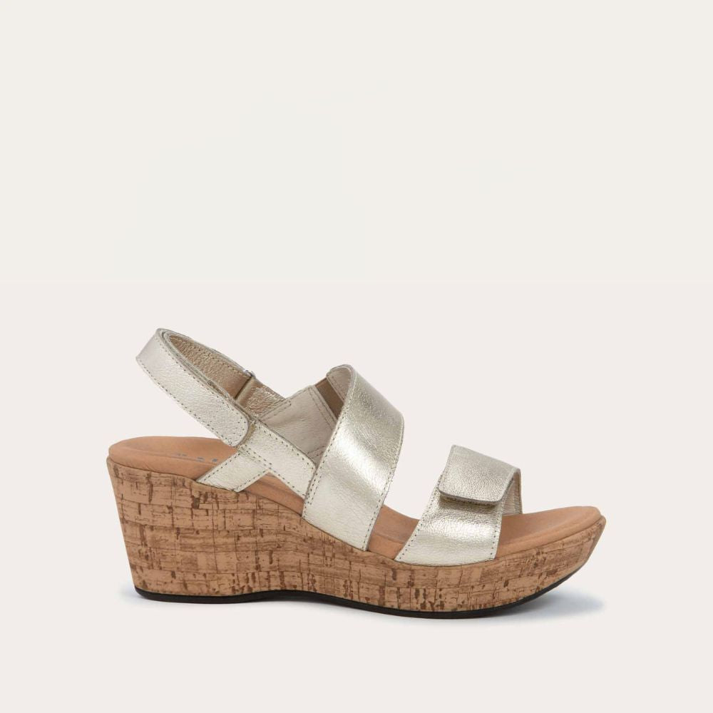 Profile view of a gold sandal with metallic straps on a cork wedge sole. Features an open toe and wedge heel.